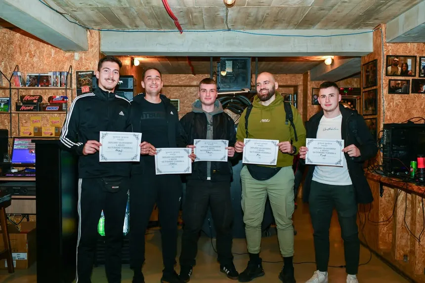 Petorica pobjednika stoje u redu i drže diplome za prvo mjesto na Turniru Turnira 4; iza njih polica sa čokoladicama, kompovi i zid s uramljenim fotografijama