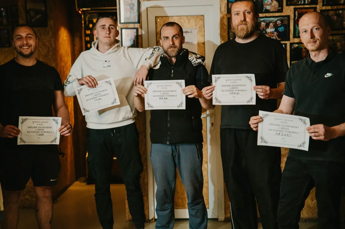 Petorica pobjednika stoje u redu u bunkeru i drže diplome za prvo mjesto na Turniru Turnira 5; iza njih bijela vrata, zidovi od OSB ploča i zid s uramljenim fotografijama