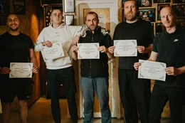 Petorica pobjednika stoje u redu u bunkeru i drže diplome za prvo mjesto na Turniru Turnira 5; iza njih bijela vrata, zidovi od OSB ploča i zid s uramljenim fotografijama