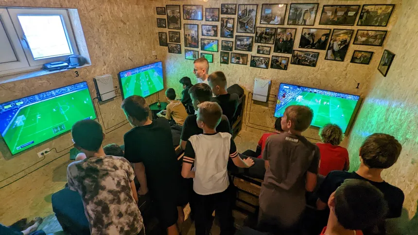 Tri ekrana uz zid, označena brojevima 1, 2 i 3 — na sva tri se igra FIFA; oko njih puna Igraona klinaca, jedni sjede na paletama, drugi stoje i gledaju