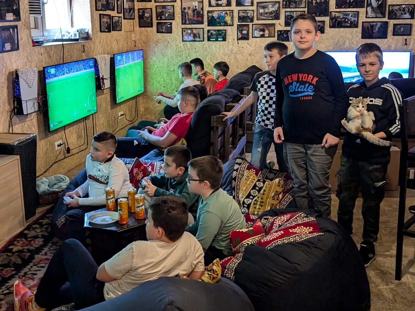 Ekipa dječaka u Igraoni — nekoliko ih igra FIFA-u na velikim ekranima, ostali sjede na vrećama ili stoje okolo; na niskom stolu limenke soka, jedan dječak drži mače, a zid iznad njih je pun uramljenih fotografija