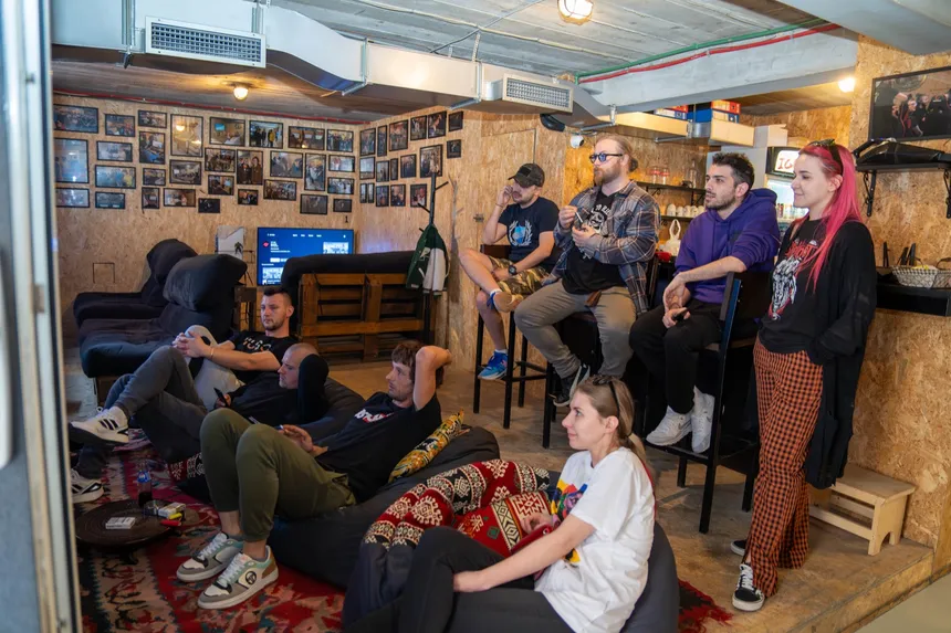 Lounge kraj šanka — nekoliko njih zavaljeno u vreće za sjedenje na ćilimu, ostali na barskim stolicama, svi okrenuti u istu stranu; iza njih zid prekriven uramljenim fotografijama